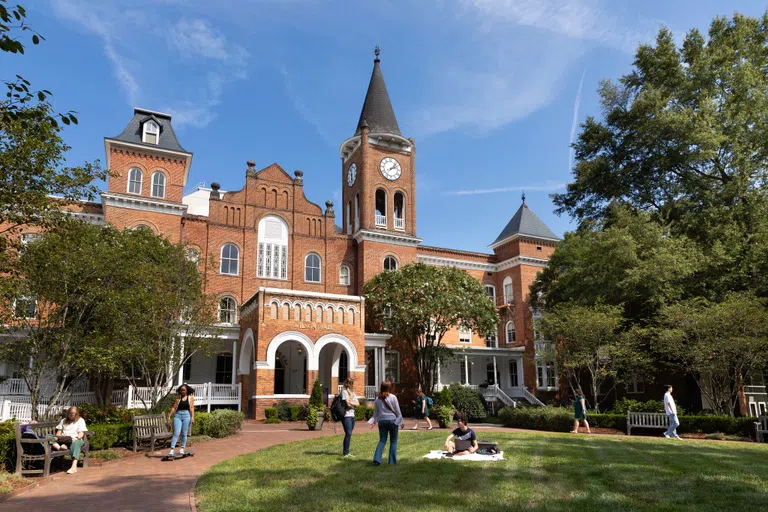 Tall brick building with four story clock tower. Green lawn with many students in foreground.