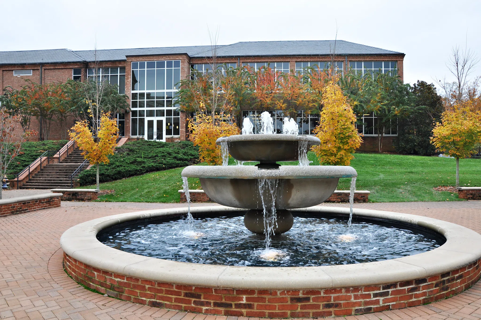 A two-tiered bronze fountain spills into a brick pool in the foreground of a two-story, brick and glass building.