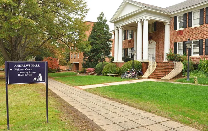Two story brick building with four-columned portico shown from angled side view. A purple sign reads: Andrews Hall; Wellness Center; Counseling Services; Health Services