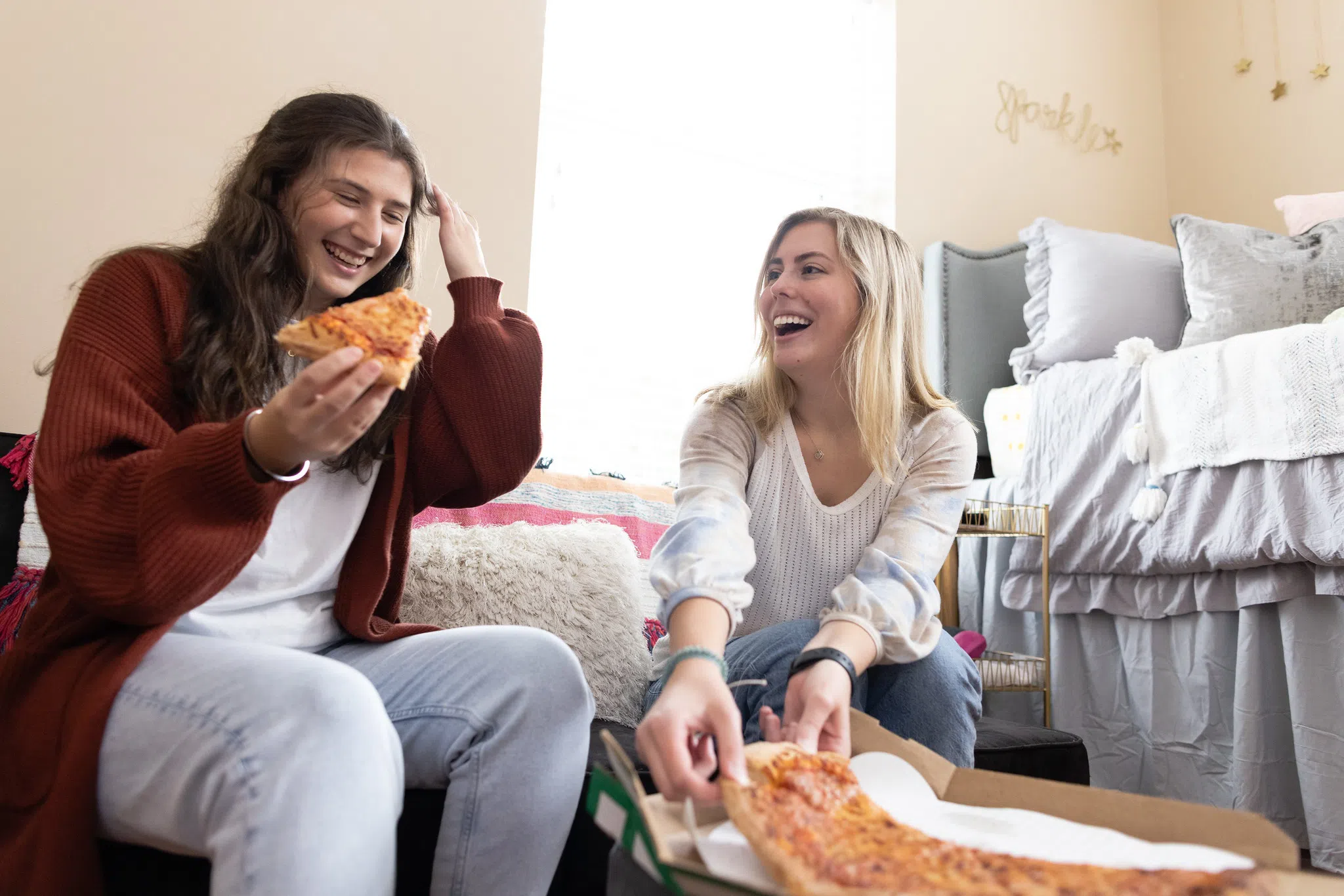 Two women laugh over a slice of pizza in a dorm room in front of a large window and a bed in the corner.