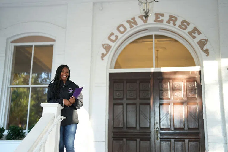 Two female students walk near wooden double doors topped by an arched window. Gold words above the arch read: CONVERSE.