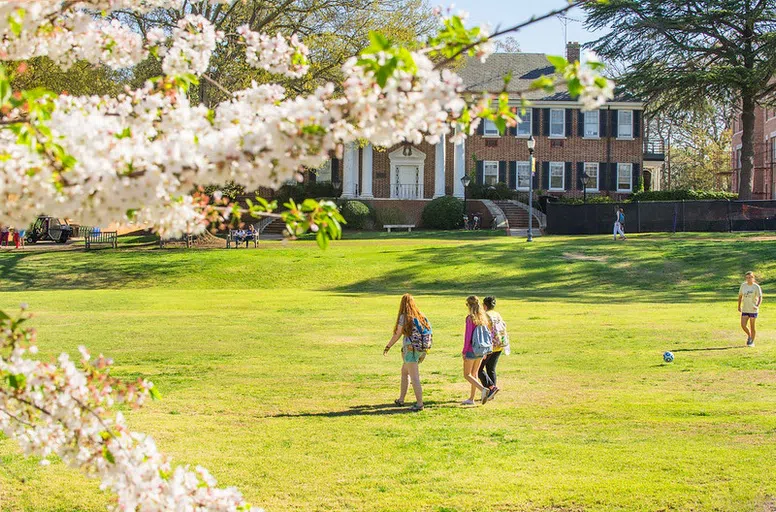 Three students walk in a grassy field in front of a two story brick building with a four-columned portico.