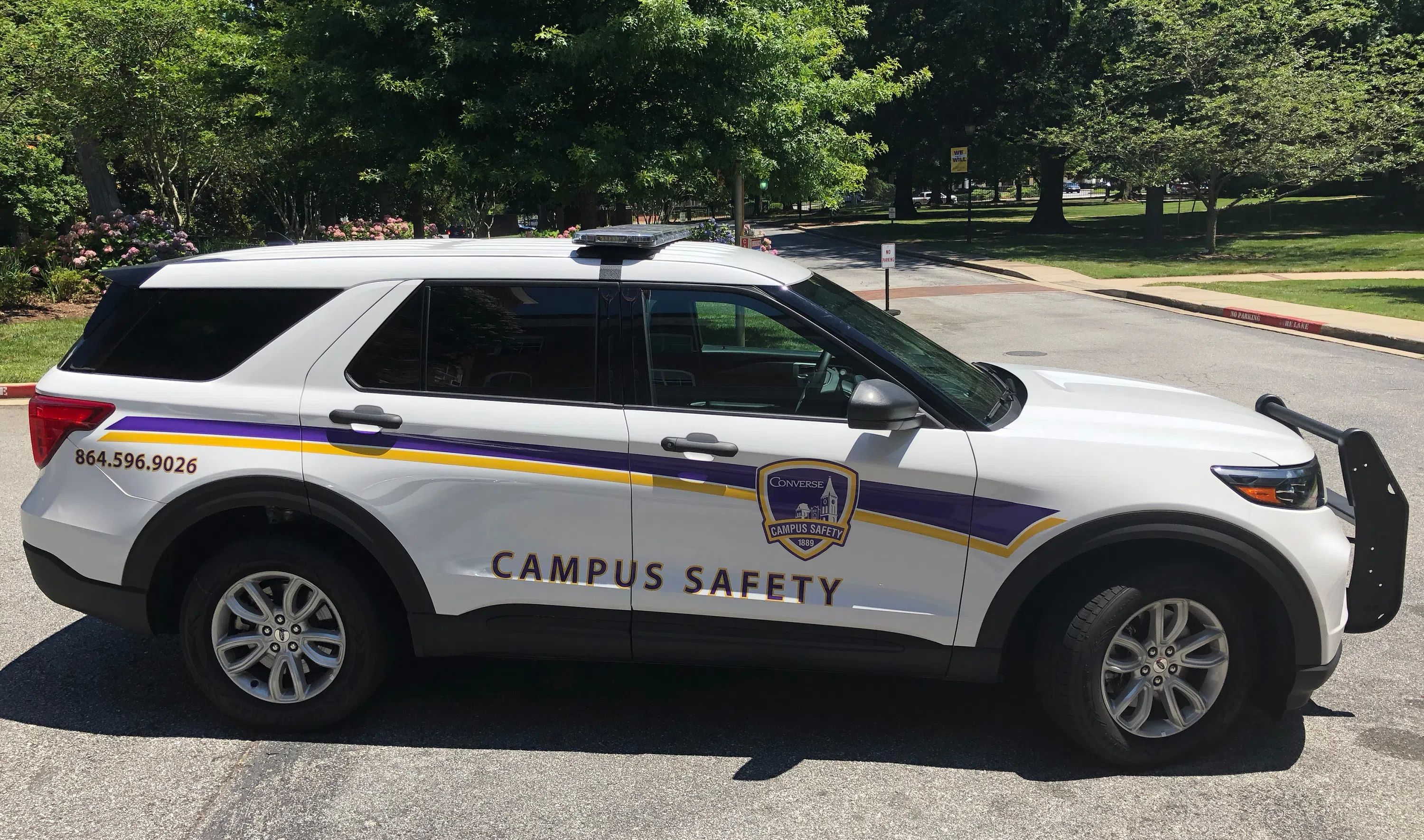 White sport utility vehicle features light bar on top and yellow and purple stripes on the side. Words on the side say "CAMPUS SAFETY" along with the phone number 864.596.9026.