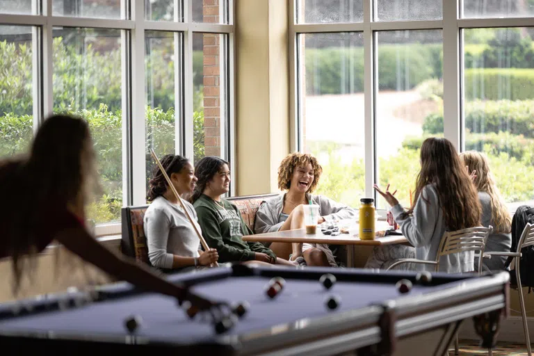 A pool table is in the foreground, and in the back students chat and laugh around a corner booth flanked by windows.