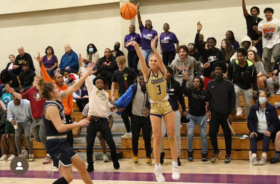 A woman in yellow Converse jersey shoots with both arms outstretched and the ball flies upward. Crowded bleachers in the background show excited fans jumping up from their seats.