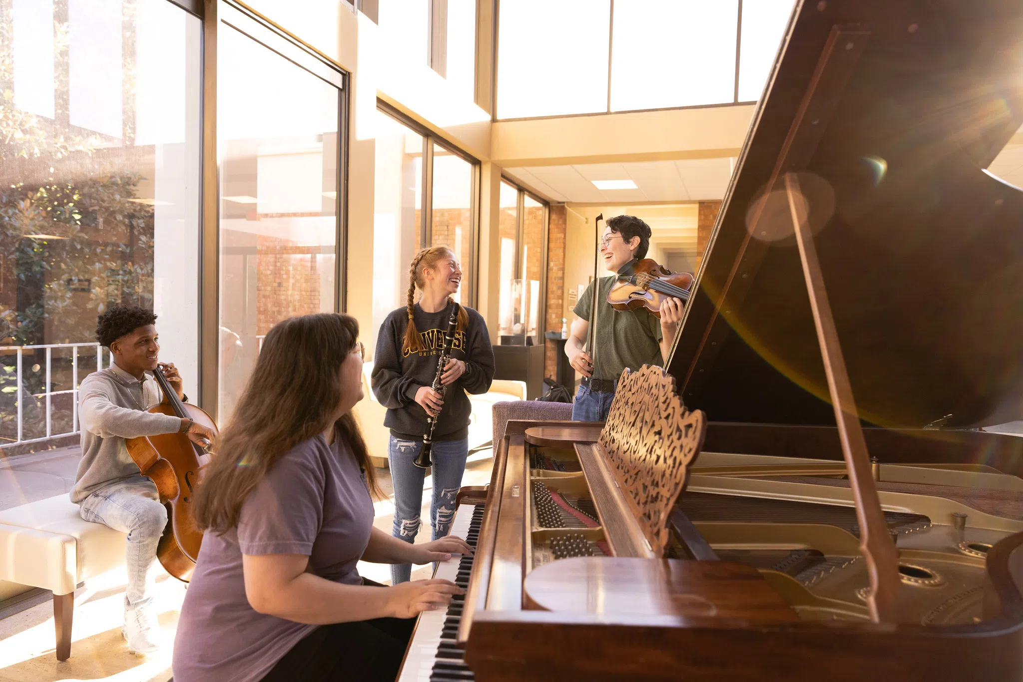 Students gather around a piano with various instruments in a sunny, window-filled room.