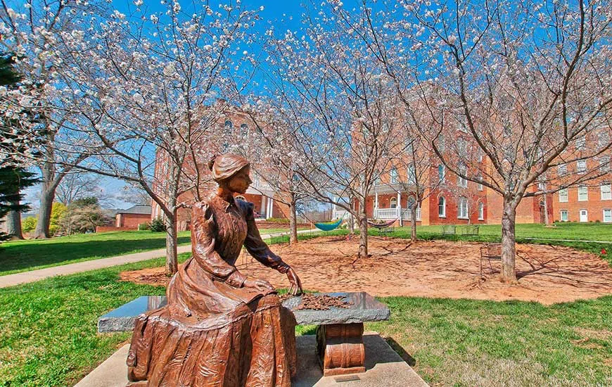 A bronze statue of a woman, Emily Dickenson, sits on a bench in an outdoor space in front of a three-story brick buidling.