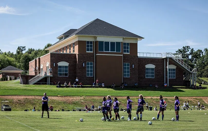 Athletes stand in a grass field below a tall brick building in sunny weather.