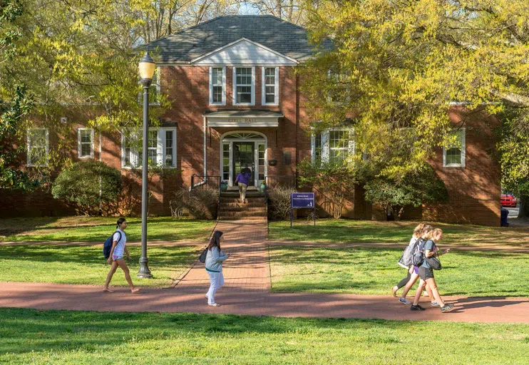 Students walk past a two story brick building with brick walkway leading up the white front door. Purple signage in front of the building marks the offices located within.