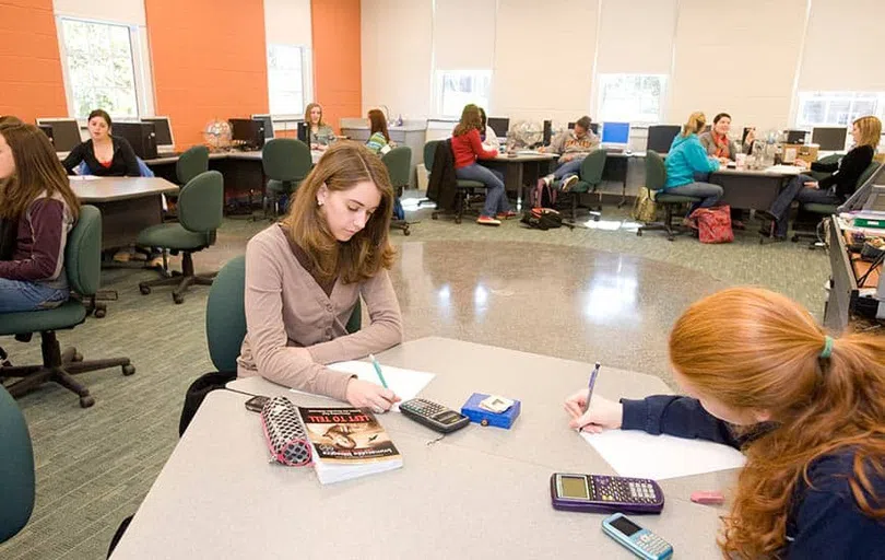 Small groups of adult students sit at work work stations surrounding a large classroom with windows and one orange accent wall.