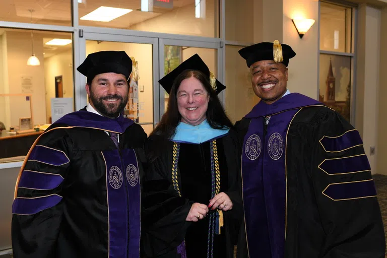 Three adult students in academic regalia smile for the camera.