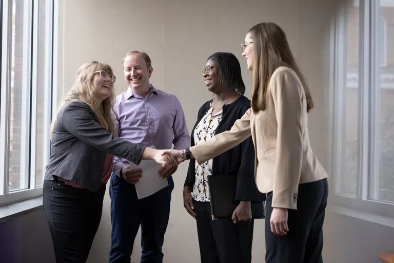 Four people smile in conversation as two shake hands with each other.