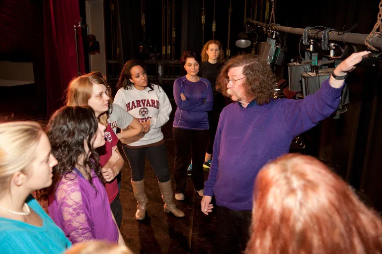 Eight individuals look at lighting supports in darkened room housing various theatre equipment.