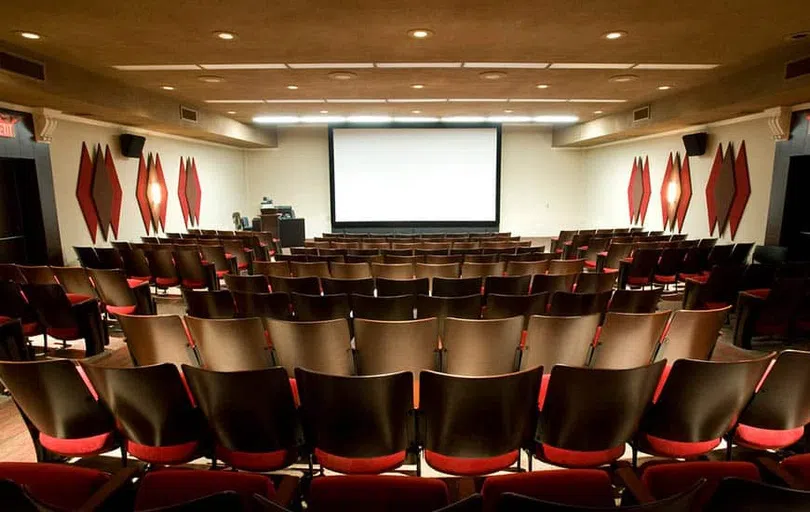 An auditorium of wooden seats with red fabric cushions face a large theatre screen.