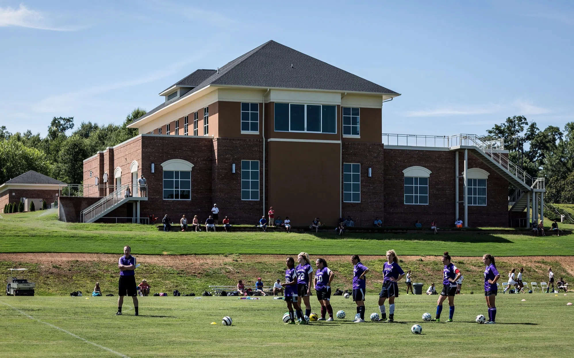 A wide brick building sits in the distance atop a green space filled with soccer players in purple jerseys.