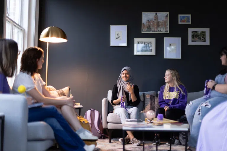 A cozy room with deep grey walls and a gallery of framed prints. Diverse women chat while seated on couches around a coffee table.