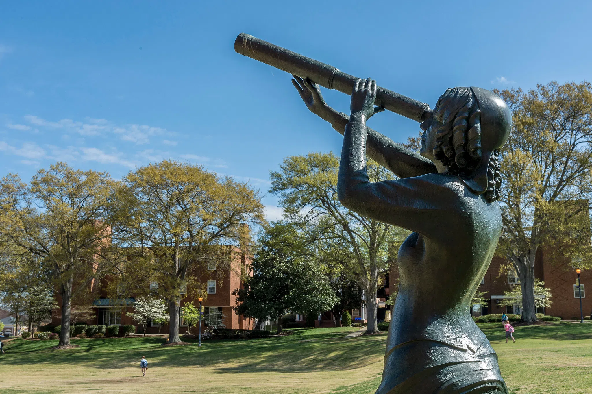 Bronze sculpture of woman with telescope appears in the foreground, with a grassy field and three-story brick building in the background.