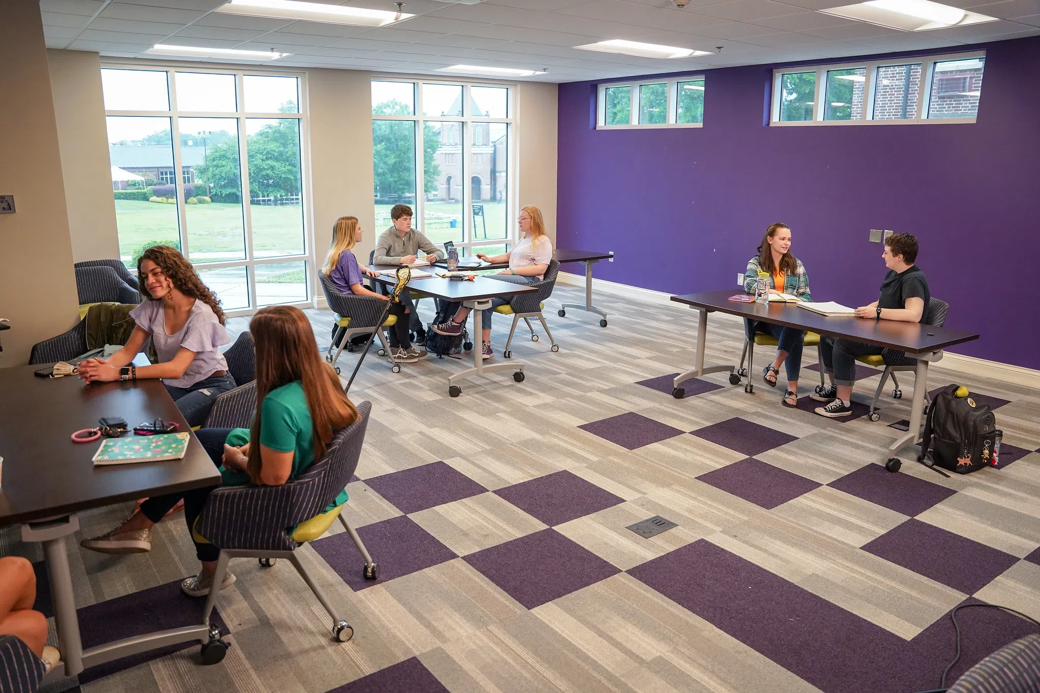 Students sit around three tables in a spacious room featuring a purple wall and windows overlooking the grassy quad.