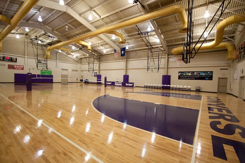 A large empty gymnasium with wood floors accented with purple zones. Yellow piping spans the ceiling.