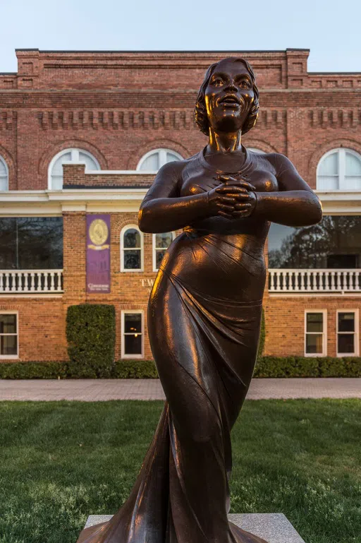 A bronze sculpture of a woman singing with her hands clasped in front of her appears in the foreground of a brick building.