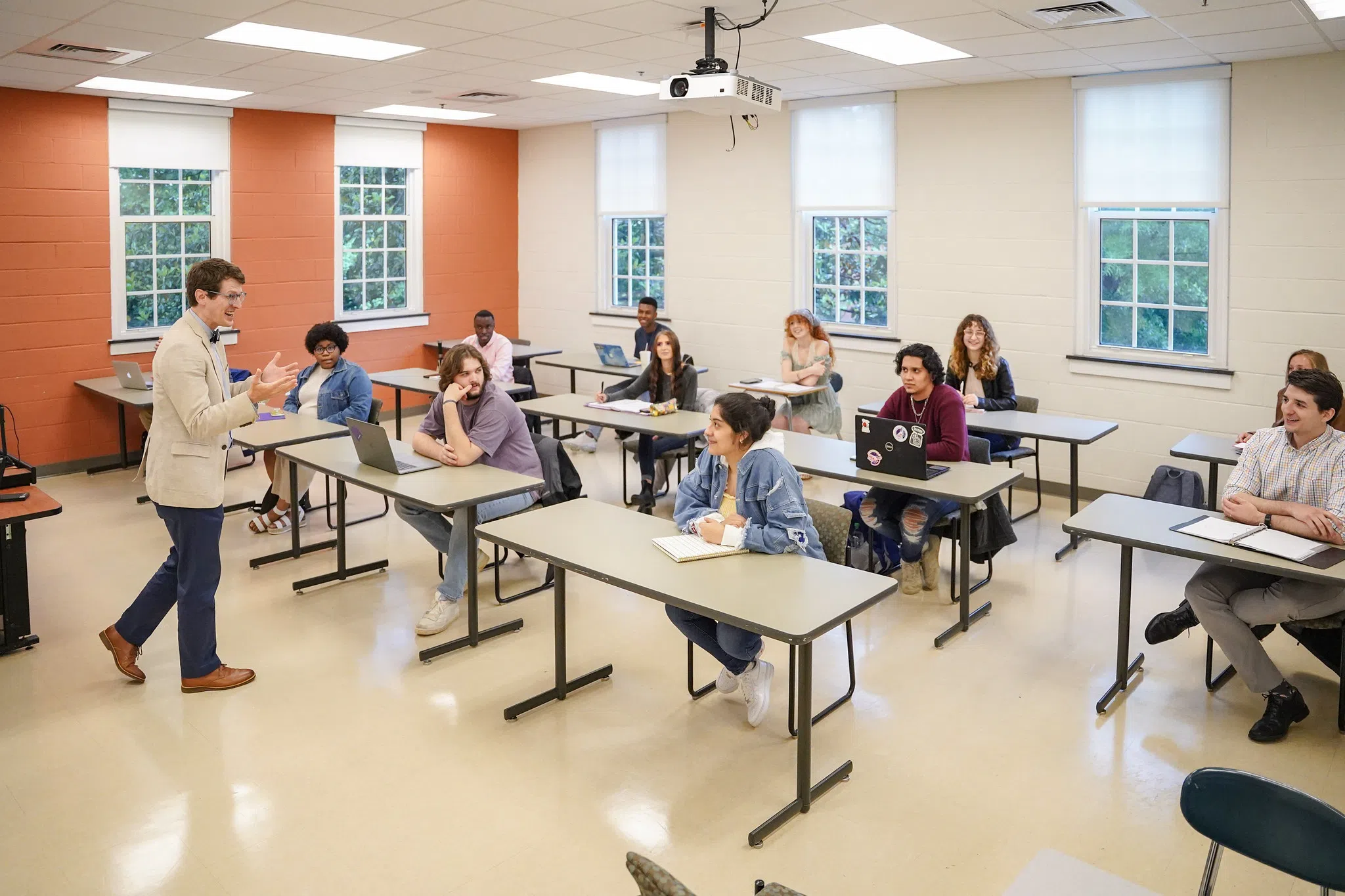 A professor on the left engages with three rows of students seated with computers out on tables.