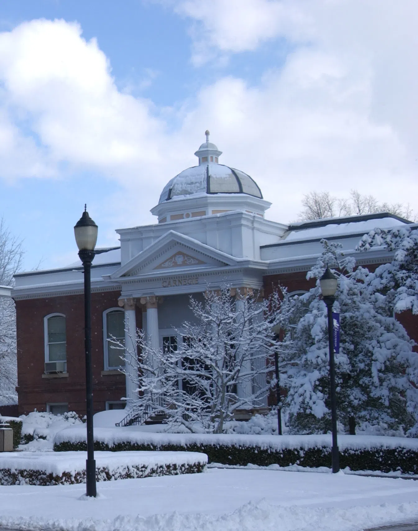 Snow covered brick building with four columns at the entrance and bronze light post in the foreground.