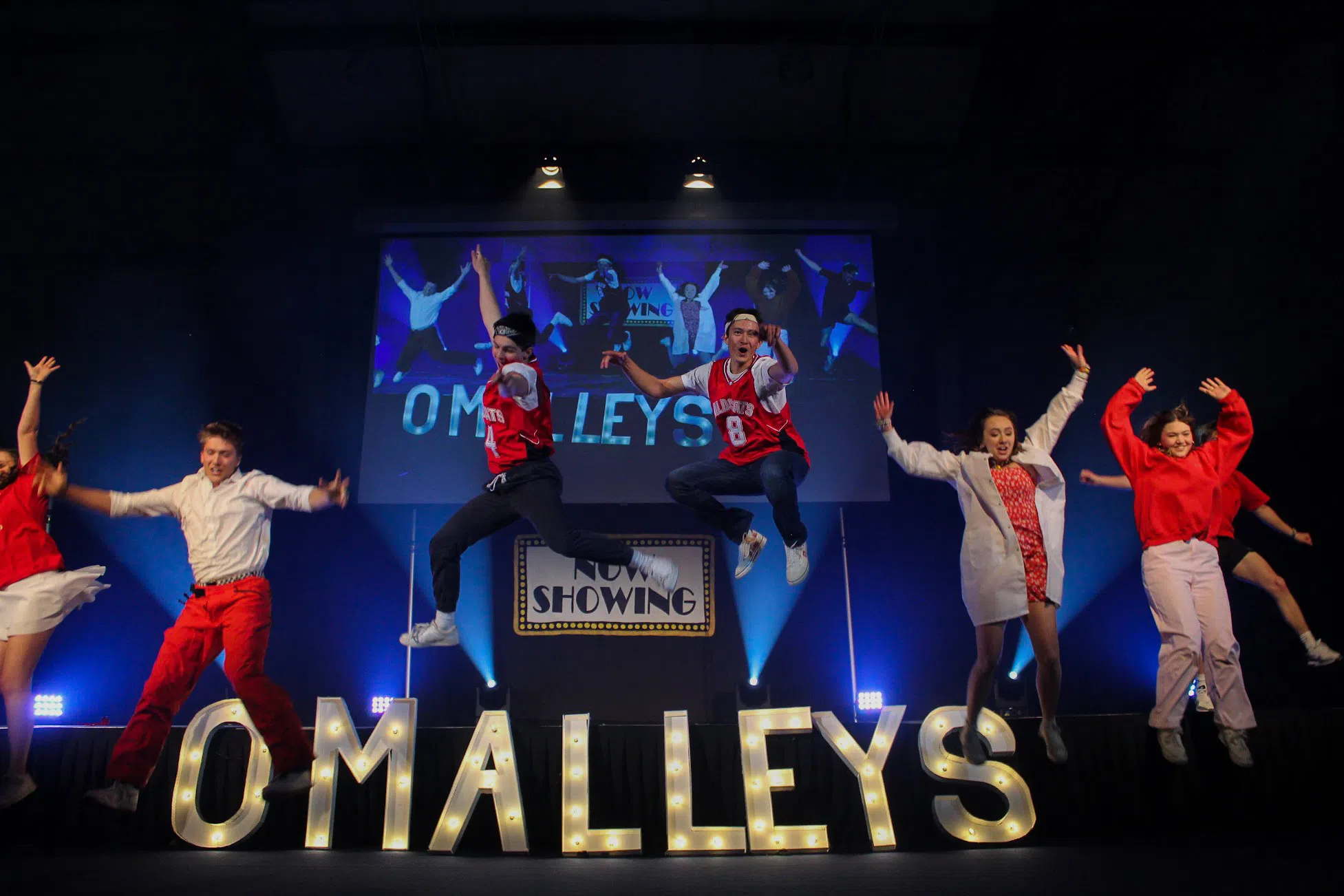 High School Musical themed cast jumping off stage with O'Malleys in bold lit up letters behind them