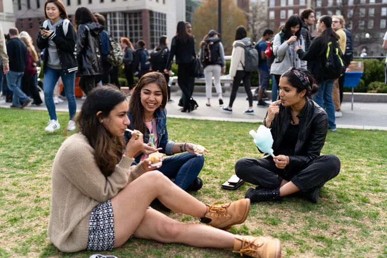 Three female students sit on the the grass in a circle while eating cotton candy and ice cream desserts.