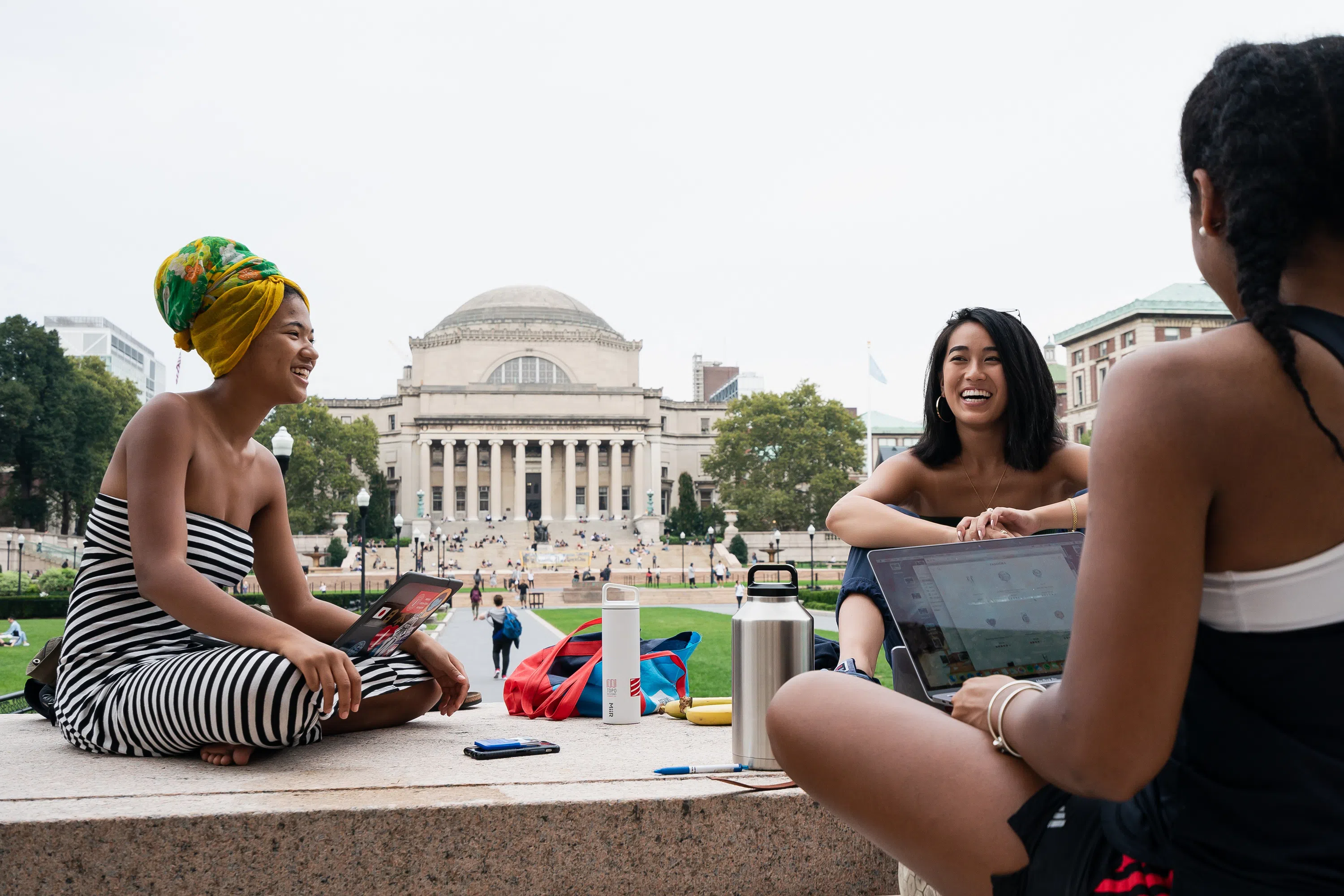 Three female students seated outside on a stone ledge, two are holding laptop computers and they are all smiling.