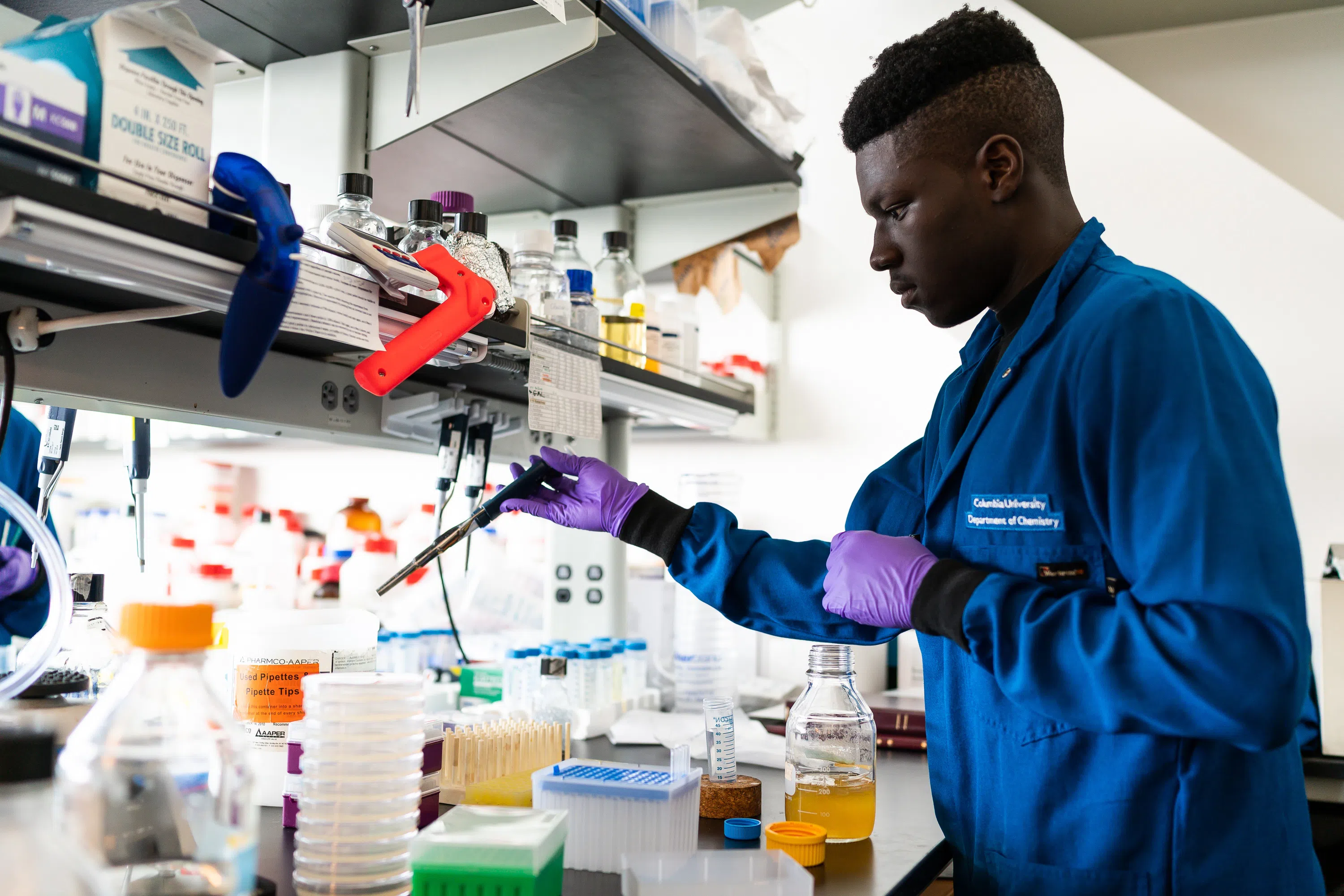 Young male student works are a lab bench, wearing a blue lab coat and pipetting materials.