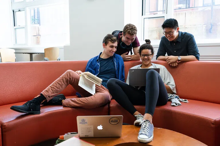 Three students sitting on red circular couch huddled a Mac laptop