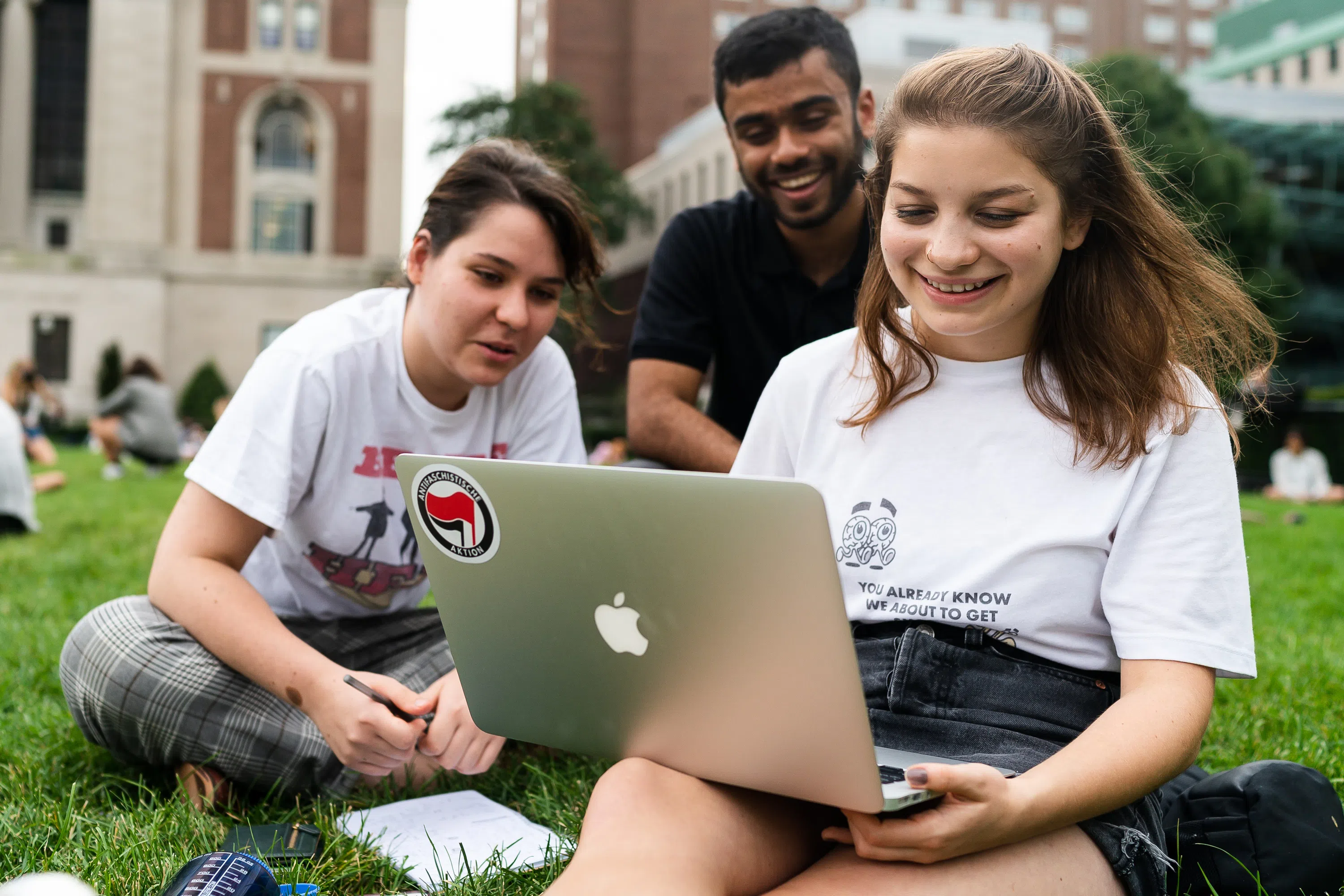 Three smiling students sitting in the grass huddled around a laptop computer