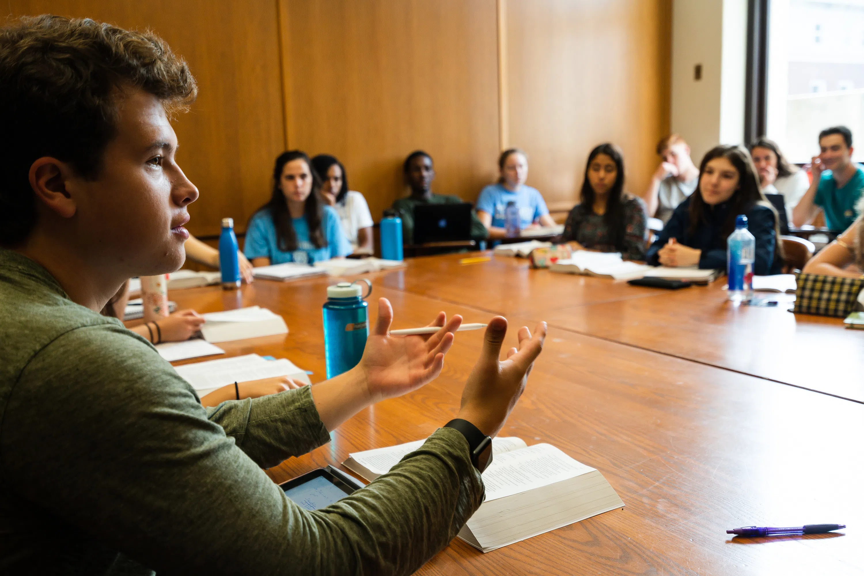 Many students sit around a large wooden table, one male student is in the foreground speaking animatedly.