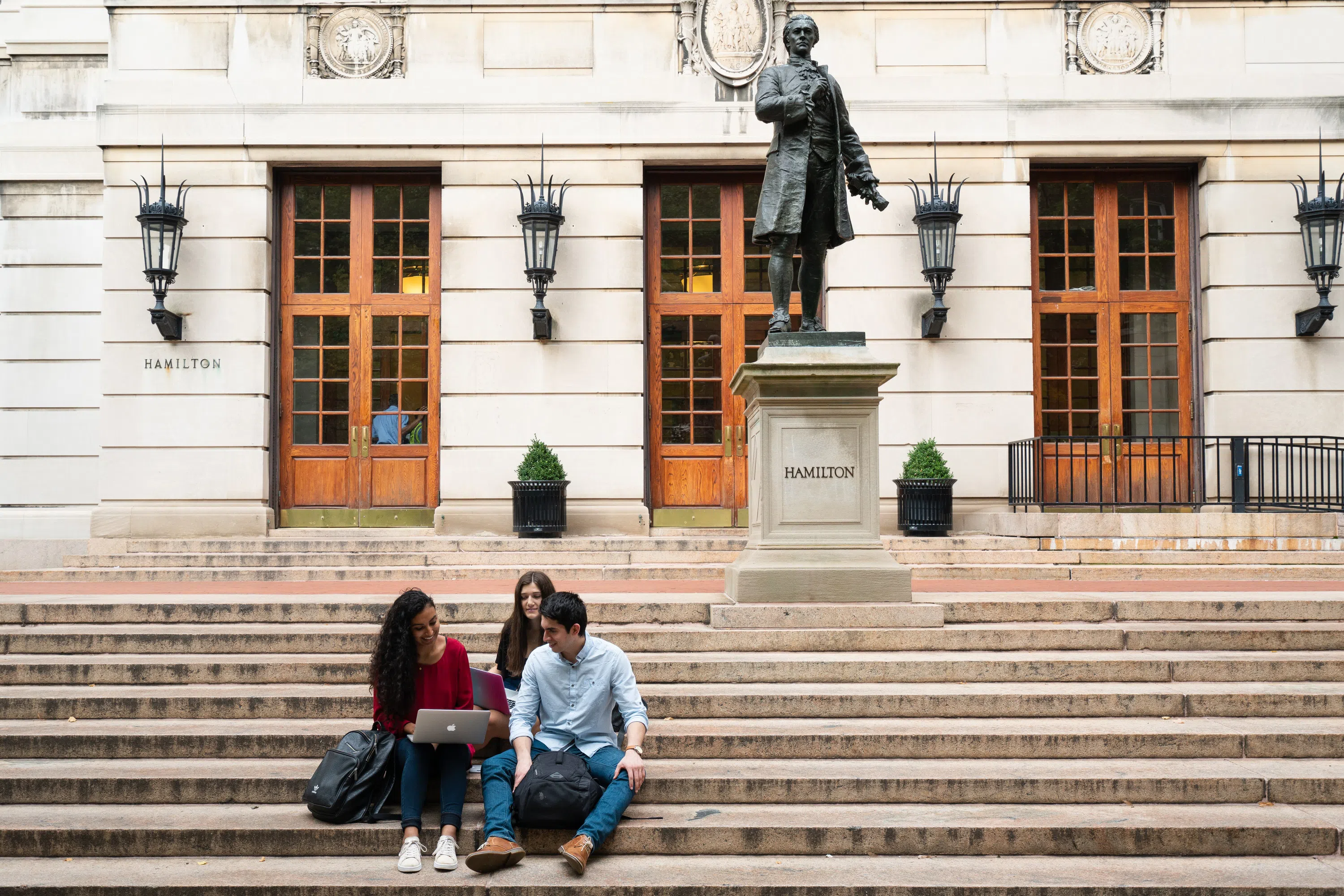 Three students sit on a set of stairs in the foreground with a bronze statue of Alexander Hamilton to the right. A classroom building sits in the background with three entryways.