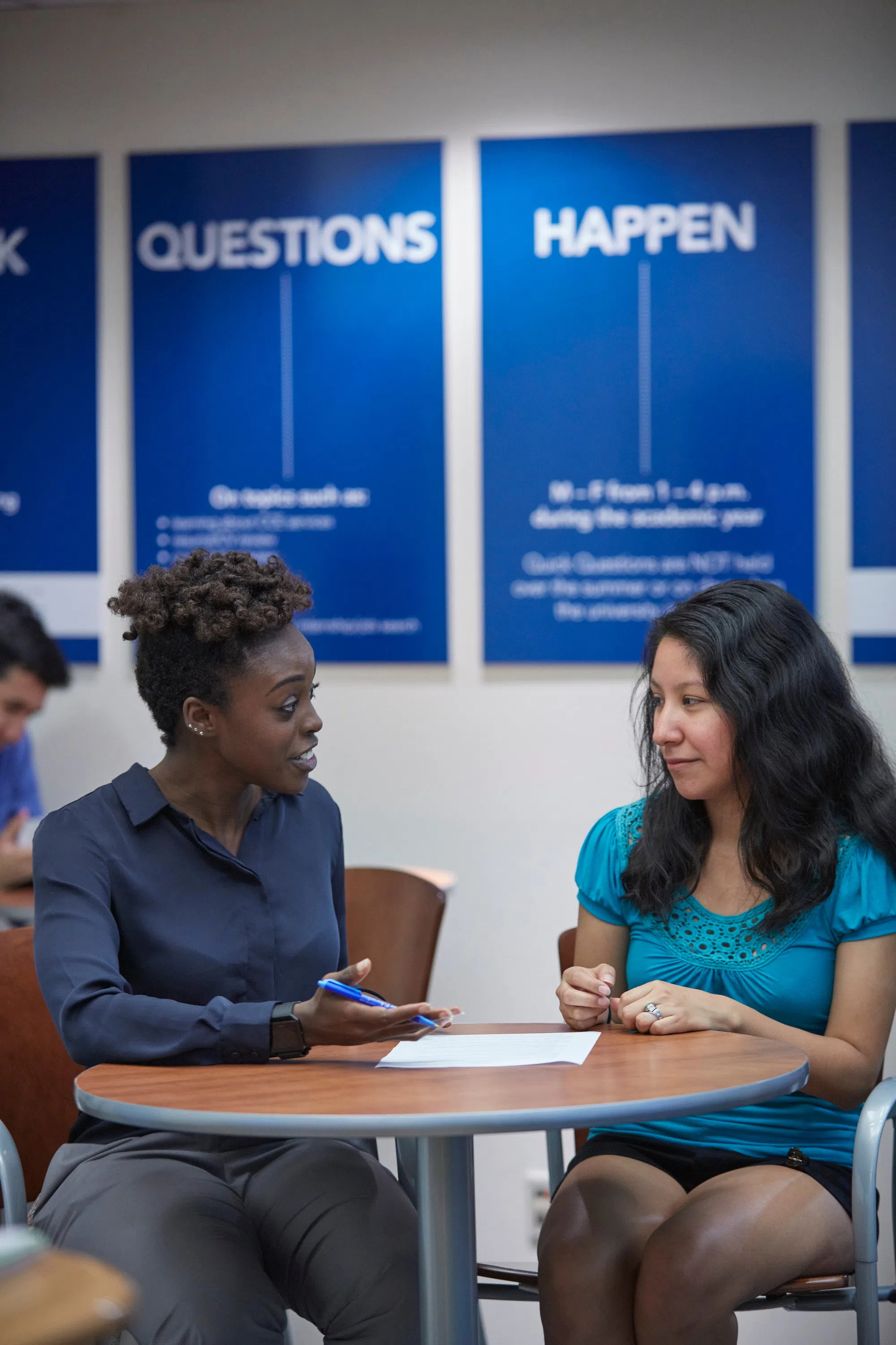 Two women sit at a small round table, there are two blue signs behind them that read "Questions Happen".