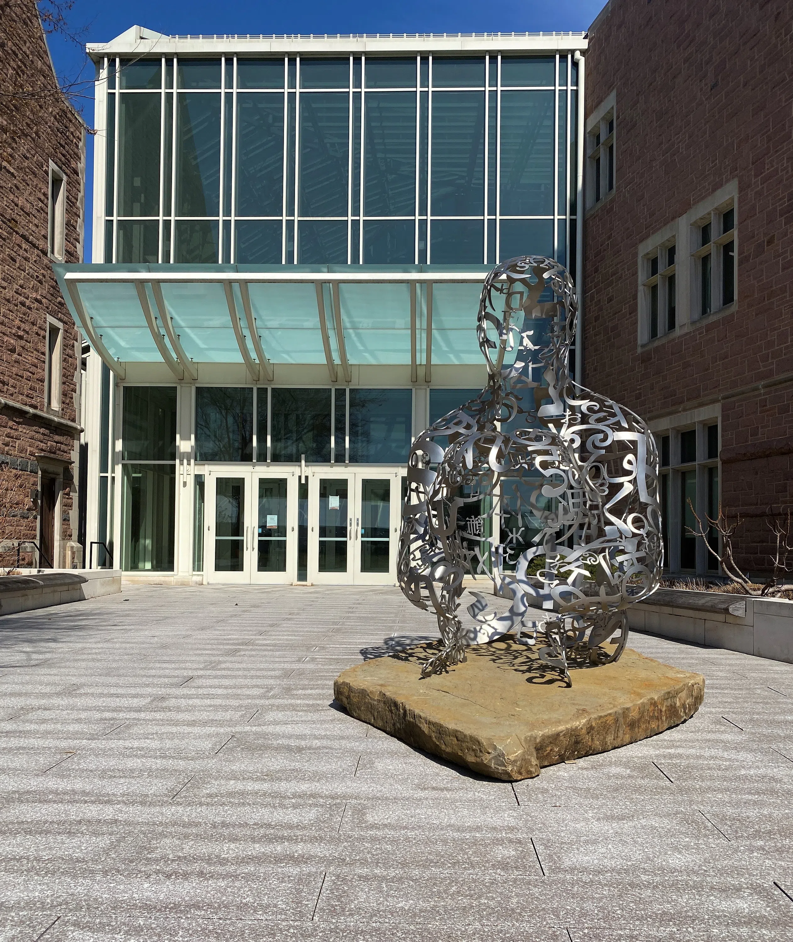 A wire-like sculpture, consisting of various numbers and symbols, of a person sitting on the ground with their knees towards their chest sits outside of Bauer Hall, directly in front of a set of glass doors.