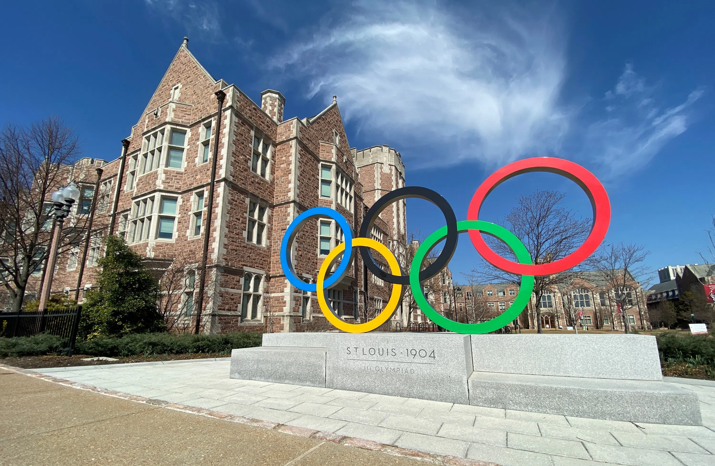 A statue of interconnected Olympic Rings, colored blue, yellow, black, green, and red, sits in front of a large, stone building and a blue sky.  The stone base of the ring sculpture reads "St. Louis 1904, III Olympiad"