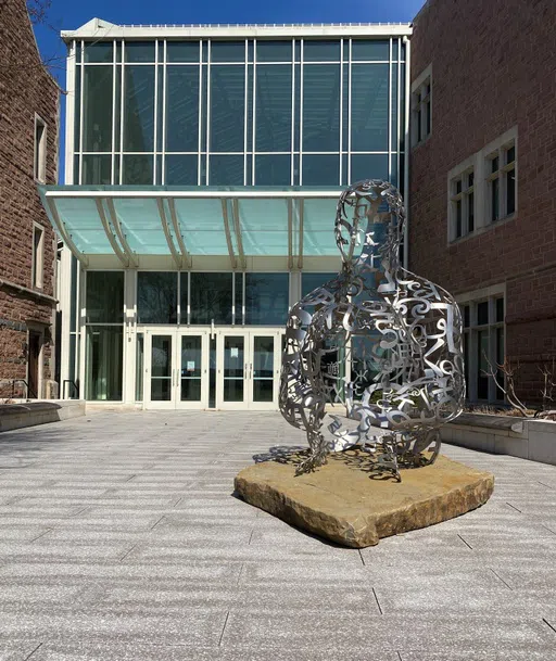 A wire-like sculpture, consisting of various numbers and symbols, of a person sitting on the ground with their knees towards their chest sits outside of Bauer Hall, directly in front of a set of glass doors.