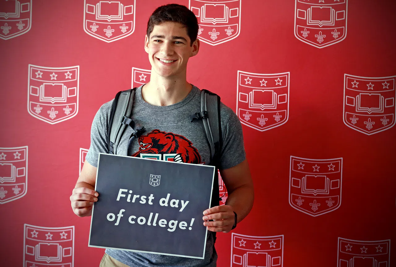 A student smiles for a photo, holding a first day of college sign