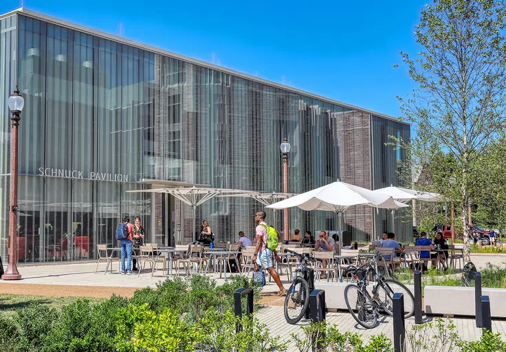 Students gather outside of a fully glass building, sitting at chairs and tables underneath a series of large patio umbrellas.