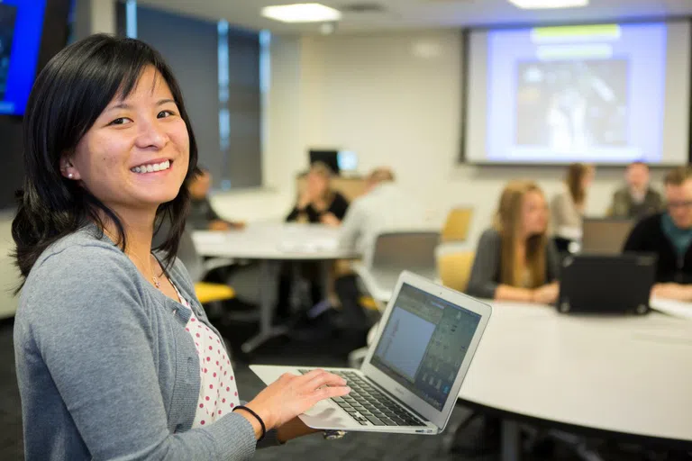 A smiling woman with a laptop in a classroom with students and a projection