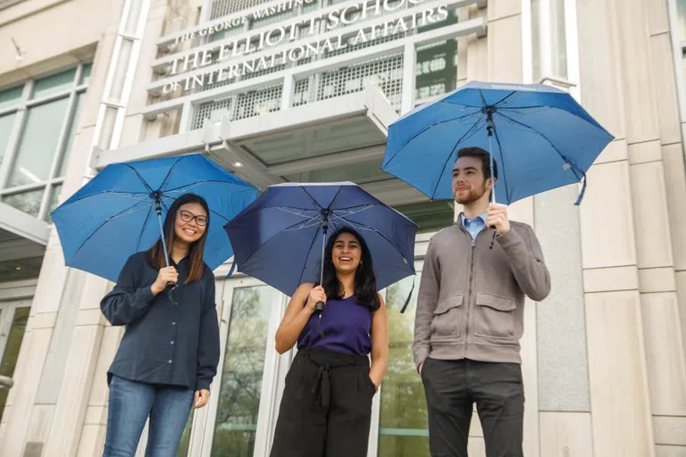 Three students with umbrellas standing in front of the Elliott School of International Affairs.