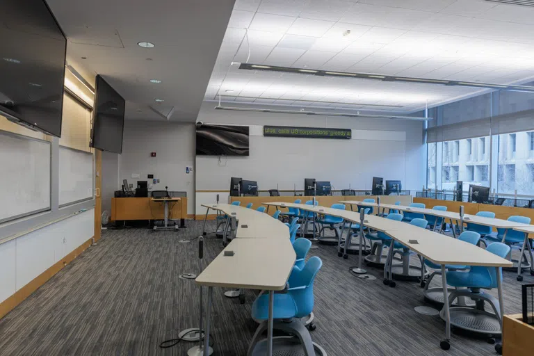 Classroom in Duquès Hall with a whiteboard, long tables and chairs. 