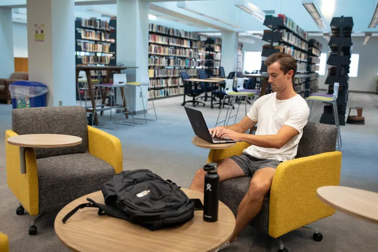 A student working on a laptop in a yellow chair with the library stacks in the background