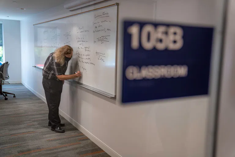 105B Classroom sign in the foreground with professor writing on the whiteboard in the background