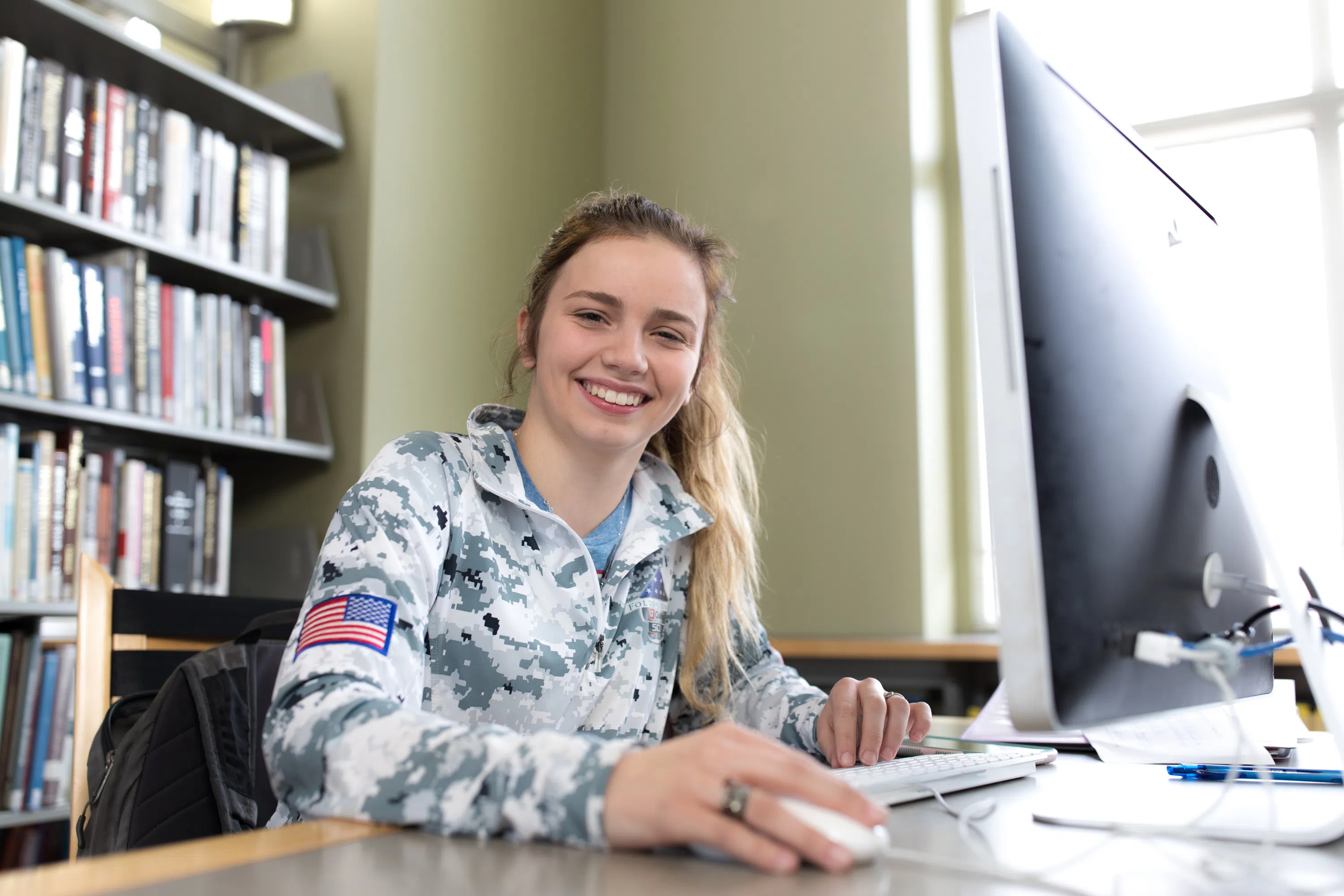 Student smiles at the camera while completing work on a desktop computer