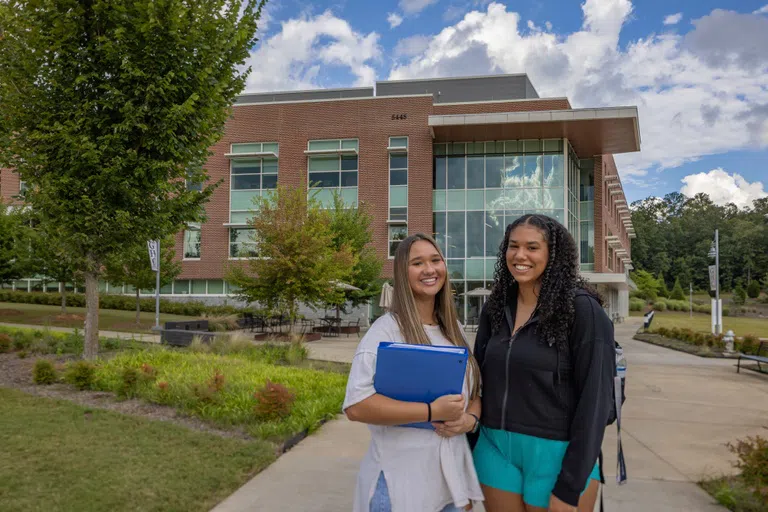 Two female students stand outside the STEAM building, smiling at the camera