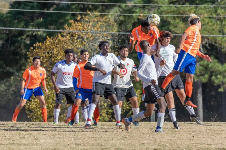 Men's soccer team, dressed in orange and blue uniforms, play against opposing team, dressed in white uniforms. 