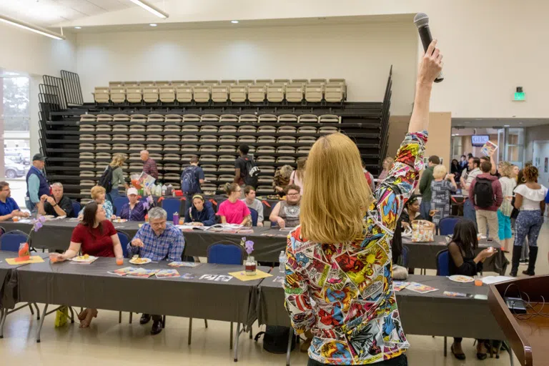 Speaker stands at the front of a group with microphone in the air. People sit in rows of tables in front of her