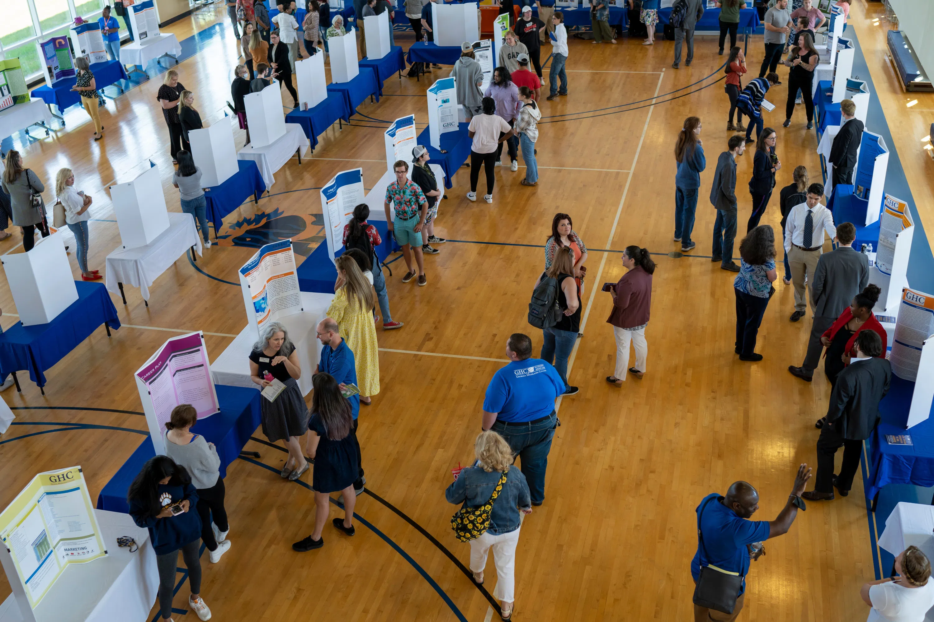 Gym converted into a fair with tables in rows and posters throughout the space. People mingle to different tables